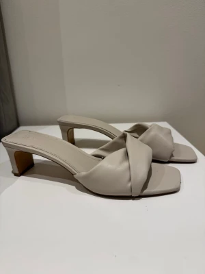 Beige slip-in sandaler med klack - Stilrena beige sandaler från H&M med låg klack och fyrkantig tå. Ovandelen har en twistad design i mjukt konstläder som ger en trendig look. Perfekta att glida i under varma dagar och passar till många outfits.