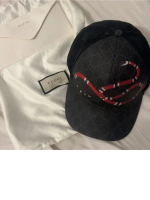 Svart Gucci keps med ormtryck - Säljer en svart keps från Gucci med det klassiska GG-mönstret och en röd, vit och svart orm broderad på framsidan. Kepsen har böjd skärm och mesh på baksidan för extra ventilation. Perfekt för dig som vill sticka ut med en lyxig accessoar.