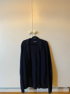 Mörkblå cardigan från Dressman - Säljer en stilren mörkblå cardiganjacka från Dressman. Cardiganen har knappar framtill, två fickor och en klassisk krage. Materialet är stickat och mjukt, perfekt för lager-på-lager. Passar dig som gillar en enkel och tidlös look.
