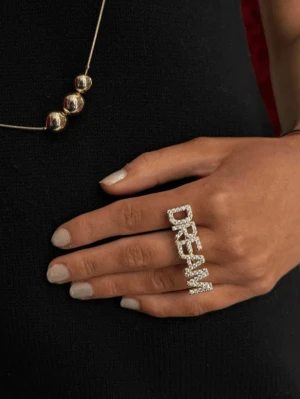 Statementring med texten DREAM - Cool statementring i silverfärg med glittrande stenar som bildar ordet DREAM. Ringen är bred och täcker två fingrar, perfekt för att sticka ut och ge din outfit en unik touch.