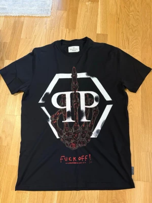 Svart t-shirt från philipp Plein med tryck - Cool svart t-shirt från Plein med stort grafiskt tryck på framsidan som visar ett skelett och texten 'fuck off!'. T-shirten har korta ärmar och är tillverkad i mjuk bomull. Perfekt för dig som gillar edgy och unika plagg. Väldigt fint skick knappt använt. Passar även L