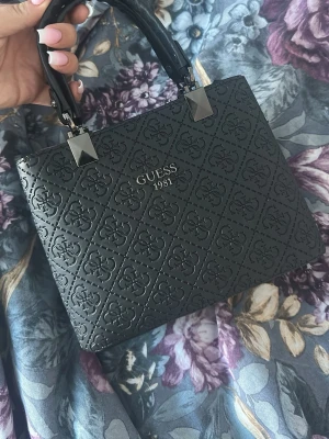  Guess - Snygg svart handväska från Guess med präglat logomönster över hela väskan och silverfärgade detaljer. Väskan har två stadiga handtag och Guess 1981 i silver på framsidan. Tillverkad i konstläder med elegant och stilren design.FAke väska 