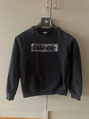 Svart tröja från Ellesse - Snygg svart tröja från Ellesse med logga i grått och svart camo på bröstet. Tröjan har rund halsringning, långa ärmar och ribbade muddar vid ärmslut och nederkant. Perfekt för en avslappnad och sportig stil.