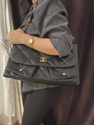 Stor svart quiltad handväska med kedja - Säljer en stor svart handväska  med guldfärgade detaljer och kedjerem. Väskan har dubbla lock, dekorativa knappar och en elegant, tidlös look. Perfekt för dig som vill ha plats för allt och samtidigt se riktigt stylish ut. Channel 