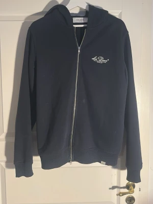 Mörkblå zip-hoodie Les Deux - Mörkblå hoodie med dragkedja från Les Deux. Klassisk design med huva, ribbade muddar och liten vit logga på bröstet. Tillverkad i mjukt bomullsmaterial som känns skönt mot huden. Perfekt för en avslappnad och stilren look.