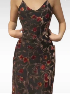Blommig långklänning med smala axelband - Snygg långklänning med blommigt mönster i rött och beige på mörk botten. Klänningen har smala axelband och v-ringad urringning, vilket ger en feminin siluett. Perfekt för dig som vill sticka ut med en elegant och trendig look.