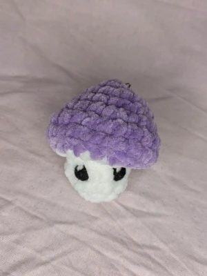 Lila och vit virkad svampnyckelring - Söt handgjord svamp i mjukt garn, virkad i lila och vitt med svarta broderade ögon. Det är också en fidget toy som är perfekt för både barn och vuxna. Perfekt som accessoar till väskan eller nyckelknippa.