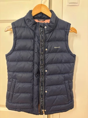 Mörkblå dunväst från GANT - Snygg mörkblå dunväst från GANT med hög krage och dragkedja med tryckknappar. Västen har två fickor fram och en ljusrosa insida som ger en cool kontrast. GANT-logga broderad på bröstet och längs dragkedjan. Perfekt för lager på lager-stil. Storlek 158/164 men passar som XS