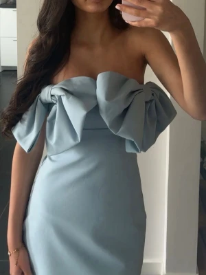 Ljusblå axelbandslös klänning med rosett - Trendig ljusblå axelbandslös klänning med stor dekorativ rosett framtill. Klänningen har en figurnära passform och är perfekt för dig som vill sticka ut. Tillverkad i ett slätt material som ger en elegant känsla.