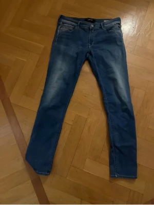 Blå slim fit jeans från Replay - Säljer ett par blå slim fit jeans från Replay med snygga slitningar och klassisk femficksmodell. Jeansen har normal midja, dragkedja och knapp i gylfen samt Replay-logga på linningen. Perfekta för en trendig och avslappnad look.