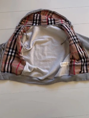 Burberry zip-up - Säljer en grå hoodie från Burberry med klassiskt rutigt foder i huvan och dragkedja framtill. Hoodien har två fickor, ribbade muddar och snören i huvan. Perfekt för dig som gillar stilrena plagg med en twist av lyx.