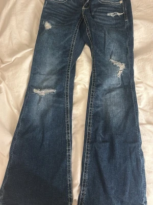 Blå slitna bootcut jeans - Snygga blå bootcut jeans med slitningar och ljusa tvättade partier. Jeansen har dekorativa sömmar på bakfickorna och en klassisk femficksmodell. Perfekta för en avslappnad och trendig look. Använt 1 gång.