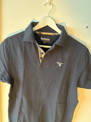 Mörkblå pikétröja  - Klassisk mörkblå pikétröja från Barbour med korta ärmar och krage. Tröjan har en liten Barbour-logga broderad på bröstet och detaljer i ljusare färg vid knäppningen. Tillverkad i mjuk bomull för en bekväm känsla.