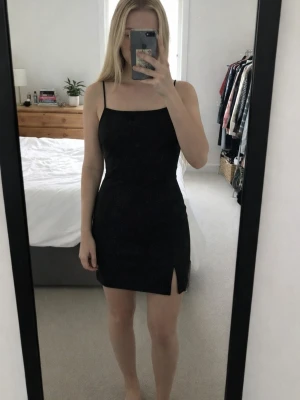 Svart glittrig kort klänning - Snygg svart klänning med glittrigt tyg och smala axelband. Modellen är kort och har en liten slits framtill. Perfekt för dig som gillar en stilren look med lite extra shimmer. Klänningen är ärmlös och har en figurnära passform.