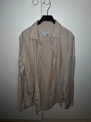 Beige skjorta från 157 med knappar - Klassisk beige skjorta från 157 med lång ärm och knappar framtill. Skjortan har en avslappnad passform och är tillverkad i mjuk bomull, perfekt för en enkel och clean look. Behövs bara en strykning