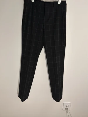 Svarta kostymbyxor rutig H&M Slim Fit - Snygga svarta kostymbyxor från H&M i slim fit-modell med vitt rutmönster. Byxorna har klassiska bälteshällor och pressveck, vilket ger en clean och modern look. Perfekta för dig som gillar stilrena och trendiga plagg.