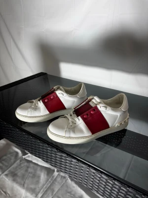 Valentino vita sneakers med röd detalj - Sick 7/10 se bild 7, skadan syns ej vid användning av skorna.                                                                        Låda och dust bag ingår i köpet. Snygga vita sneakers från Valentino med bred röd läderdetalj över snörningen och coola nitar på hälen. Skorna har en klassisk låg profil och är tillverkade i läder. Perfekt för dig som vill sticka ut med stilrena och exklusiva sneakers.