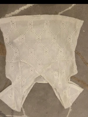 Vit spetsig omlott topp - Säljer en vit omlott topp med spetsdetaljer och broderat hålmönster. Toppen är ärmlös och har en unik form med omlott-design framtill. Perfekt för dig som gillar romantiska och feminina plagg.