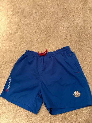 Blå badbyxor från Moncler - Snygga blå badbyxor från Moncler med röd dragsko i midjan och Moncler-logga på vänster ben. Klassisk modell med elastisk midja och diskreta röda detaljer vid fickorna. Perfekta för stranden eller poolen.