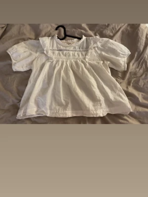 Vit blus med spetsdetaljer - Söt vit blus med korta puffärmar och volangdetaljer vid axlarna. Blusen har en romantisk känsla med spets och broderade detaljer framtill. Tillverkad i ett lätt tyg som ger en luftig och avslappnad passform.