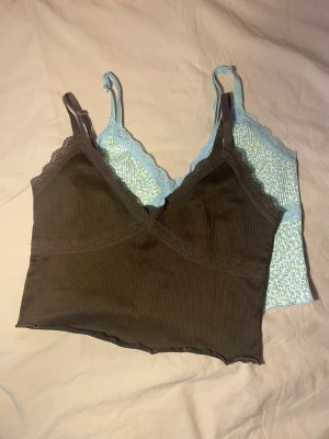 Två ribbade linnetoppar med spets set  - Säljer två söta linnetoppar, en brun ribbad med spetsdetaljer och en ljusblå med småblommigt mönster och spetskant. Båda har smala axelband och croppad passform, perfekta för lager-på-lager eller sommaroutfits. De har viss tecken på användning, till exempel noppror.