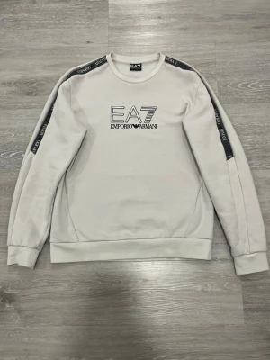 Vit stickad tröja från Emporio Armani - Snygg vit stickad tröja från Emporio Armani EA7 med svart logga på bröstet och svarta band med text längs axlarna. Tröjan har rund halsringning, ribbade muddar och en avslappnad passform. Perfekt för dig som gillar stilrena och exklusiva plagg.