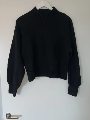 Svart stickad tröja med hög hals - En svart stickad tröja med hög ribbad hals och breda muddar vid ärmslut och nederkant. Tröjan har en rak och något boxig passform med långa ärmar.