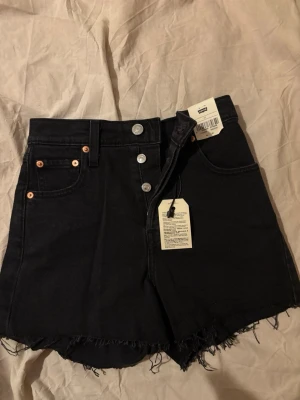 Svarta shorts från Levi's - Snygga svarta high waist shorts från Levi's med rå kant nertill och klassisk femficksdesign. Knappar framtill i silver och Levi's ikoniska läderpatch bak. 