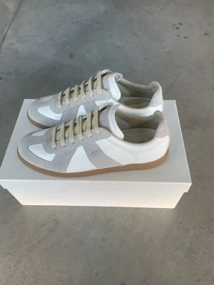 Maison margiela gats  - Snygga sneakers i vitt och ljusgrått med beige mockadetaljer och klassisk gummisula. Skorna har vita och grå paneler, beige snörning och en stilren retrodesign som passar till det mesta. Perfekta för dig som gillar minimalistisk och clean stil.