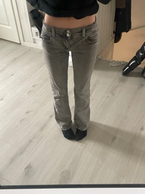 Grå bootcut jeans med låg midja - Säljer ett par gråa bootcut jeans med låg midja och klassiska fem fickor. Jeansen har en snygg tvättad look och stängs med dragkedja och knapp. Perfekta för dig som gillar en avslappnad men trendig stil.