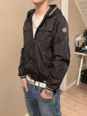Svart Moncler vindjacka - Snygg svart vindjacka från Moncler med logotyp på ärmen. Jackan har två bröstfickor med knapp och två sidofickor. Passar perfekt nu till våren och hela sommaren. Qr koden tar dig till code moncler istället för deras hemsida (de som är fejk tar än till hemsidan istället för code). Pris går att diskutera!