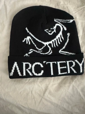 Svart mössa från Arc'teryx - Snygg svart mössa från Arc'teryx med vit logga och coolt skelettmönster framtill. Mössan är stickad och har uppvikt kant med tydlig text. Perfekt för dig som gillar streetwear och outdoor vibes.