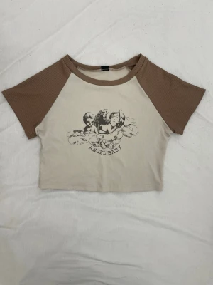 Beige och brun croppad t-shirt Angel Baby - Trendig croppad t-shirt med beige kropp och bruna ärmar. Framsidan har ett svart tryck med änglar och texten 'ANGEL BABY'. T-shirten har korta ärmar och är tillverkad i mjuk bomull, perfekt för en avslappnad stil.