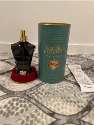 Jean Paul Gaultier Le Beau Le Parfum - Unik parfymflaska formad som en svart manlig torso med gulddetaljer och spraymunstycke. Kommer i en stilren grön metalltub med guldfärgad text och lock. Flaskan står på en röd sammetsliknande bas. Perfekt för dig som gillar exklusiva dofter och snygg design.