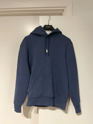 Blå hoodie från Polo Ralph Lauren - Snygg blå hoodie från Polo Ralph Lauren med klassisk huva och dragsko. Har en vit broderad logga på bröstet och stor känguruficka framtill. Tillverkad i mjukt bomullsmaterial och har ribbade muddar vid ärmar och nederkant. Perfekt för en chill och stilren look.