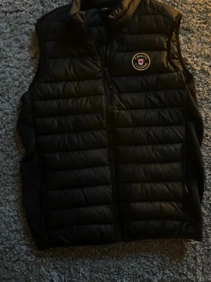 Svart väst från Zavetti Canada - Snygg svart dunväst från Zavetti Canada med quiltad design och dragkedja framtill. Västen har en hög krage och ett broderat märke på bröstet. Perfekt för lager-på-lager och passar till många olika outfits. Priset är diskuterbart!