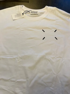 Vit Maison Margiela T-shirt - Säljer en vit T-shirt från Maison Margiela med minimalistiskt tryck framtill och stort grafiskt tryck med logga på ryggen. Klassisk rund hals och korta ärmar. Tillverkad i mjuk bomull för en clean och stilren look.