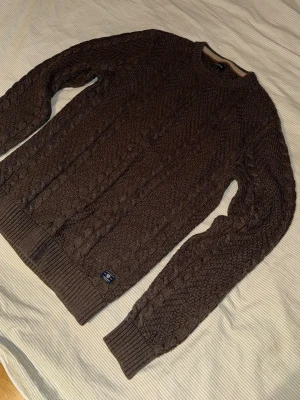 Brun kabelstickad tröja - Jack&Jones  - Säljer en brun kabelstickad tröja från Jack&Jones med rund hals och ribbade muddar vid ärmslut och nederkant. Använt skick men fint i övrigt. Tröjan har ett klassiskt mönster och är perfekt för lager-på-lager. Märkeslapp sitter på nederkanten och i nacken.
