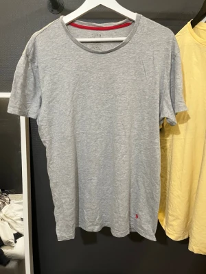 Grå t-shirt från Polo Ralph Lauren - Snygg grå t-shirt från Polo Ralph Lauren med klassisk röd broderad logga på nedre delen. T-shirten har rund halsringning och korta ärmar. Tillverkad i mjuk bomull som känns skön mot huden. Perfekt basplagg till garderoben.