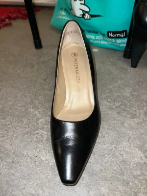 Svarta pumps från Peter Kaiser - Stilrena svarta pumps från Peter Kaiser i slätt läder. Skorna har spetsig tå och medelhög klack som ger en elegant siluett. Insidan är beige och skon har en klassisk, tidlös design som passar till många olika looks.