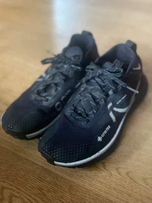 Nike svart Gore-Tex löparsko - Svarta löparskor från Nike med Gore-Tex-material som gör dem vattentäta. Skorna har vita detaljer, snörning och en slitstark sula för bra grepp. Perfekta för löpning på olika underlag och när du vill hålla dig torr och bekväm.