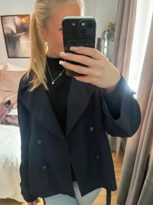 Mörkblå trenchcoat med knytskärp - Säljer en stilren mörkblå trenchcoat med breda slag, dubbelknäppning och knytskärp i midjan. Jackan har en klassisk och rak passform, perfekt för att ge en snygg siluett. Materialet känns mjukt och lätt, vilket gör den bekväm att bära.