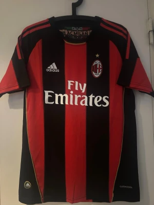 AC Milan matchtröja Adidas röd/svart - Säljer en klassisk AC Milan matchtröja från Adidas med breda röda och svarta ränder, klubbmärke och Fly Emirates-tryck på bröstet. Tröjan har korta ärmar och är tillverkad i lätt och ventilerande Climacool-material. Perfekt för fotbollsträning eller att bära på läktaren.