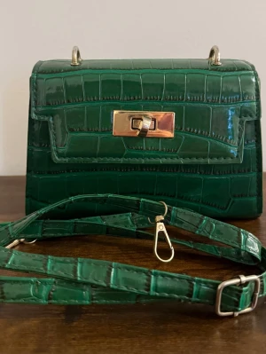 Grön krokopräglad handväska - Stilren grön handväska i krokopräglat konstläder med guldfärgade metalldetaljer och vridlås. Väskan har en avtagbar och justerbar axelrem i samma material och färg. Perfekt accessoar för att lyfta din outfit med en trendig touch.