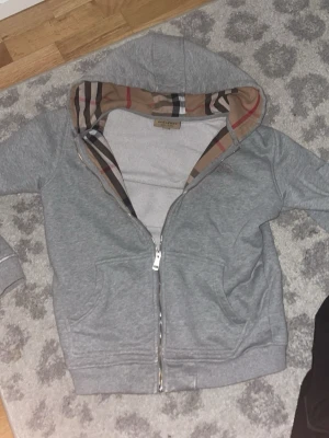 Grå hoodie från Burberry med dragkedja - Grå hoodie från Burberry med klassiskt rutigt foder i huvan och dragkedja framtill. Hoodien har två fickor och en broderad Burberry-logga på bröstet. Mjuk bomullsblandning och ribbade muddar för extra komfort.