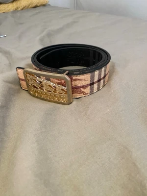 Beige-randigt bälte med gulddetalj - Snyggt bälte från Burberry med klassiskt beige och brunt rutmönster samt en stor guldfärgad metallspänne med logga. Bältet har svarta kanter och känns riktigt lyxigt. Perfekt accessoar för att lyfta din outfit.