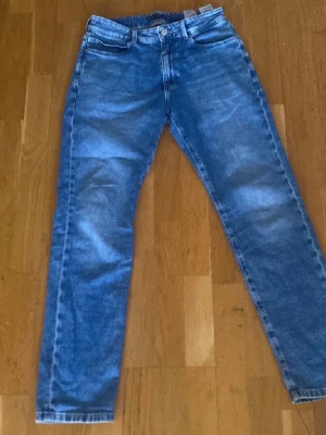 Blå raka jeans från Tommy Hilfiger - Snygga blå jeans från Tommy Hilfiger med klassisk femficksmodell och raka ben. Jeansen har en ljus tvätt och detaljer som läderpatch med logga samt den ikoniska flaggan på bakfickan. Perfekta för en avslappnad och trendig stil.