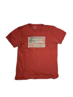 Röd t-shirt från Ralph Lauren - Röd t-shirt från Ralph Lauren med amerikansk flagga och logga tryckt på bröstet. Klassisk passform med korta ärmar och rund halsringning. Tillverkad i mjuk bomull som känns skön mot huden. Perfekt för en avslappnad och stilren look.