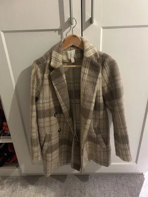 Beige-rutig ullkappa med dubbelknäppning - Snygg beige och ljusbrun rutig kappa med dubbelknäppning och klassisk krage. Kappan har två fickor framtill och är tillverkad i ett mjukt ullblandat material. Perfekt för dig som vill ha en trendig och tidlös look i höst och vinter.