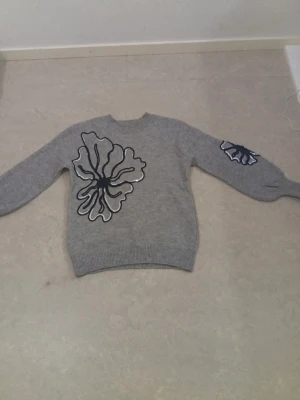 Grå stickad tröja med blommor - Grå stickad tröja med stora blommiga applikationer i svart och vitt på bröstet och ena ärmen. Tröjan har rund halsringning, långa ärmar och ribbstickade muddar vid ärmslut och nederkant. Perfekt för dig som gillar unika detaljer.